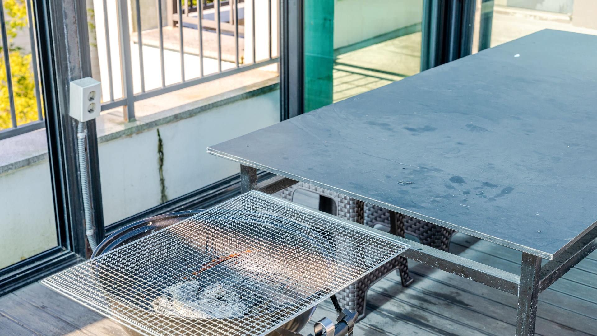 Et bord med metalrist og en netkurv nedenunder, placeret på en træterrasse med en glasdør i baggrunden.