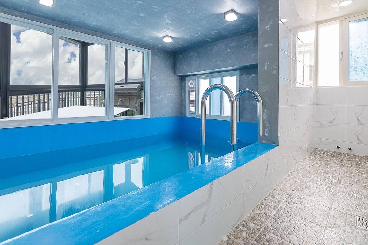 A swimming pool with a blue edge, a shower area, and a view of the outdoors through large windows.