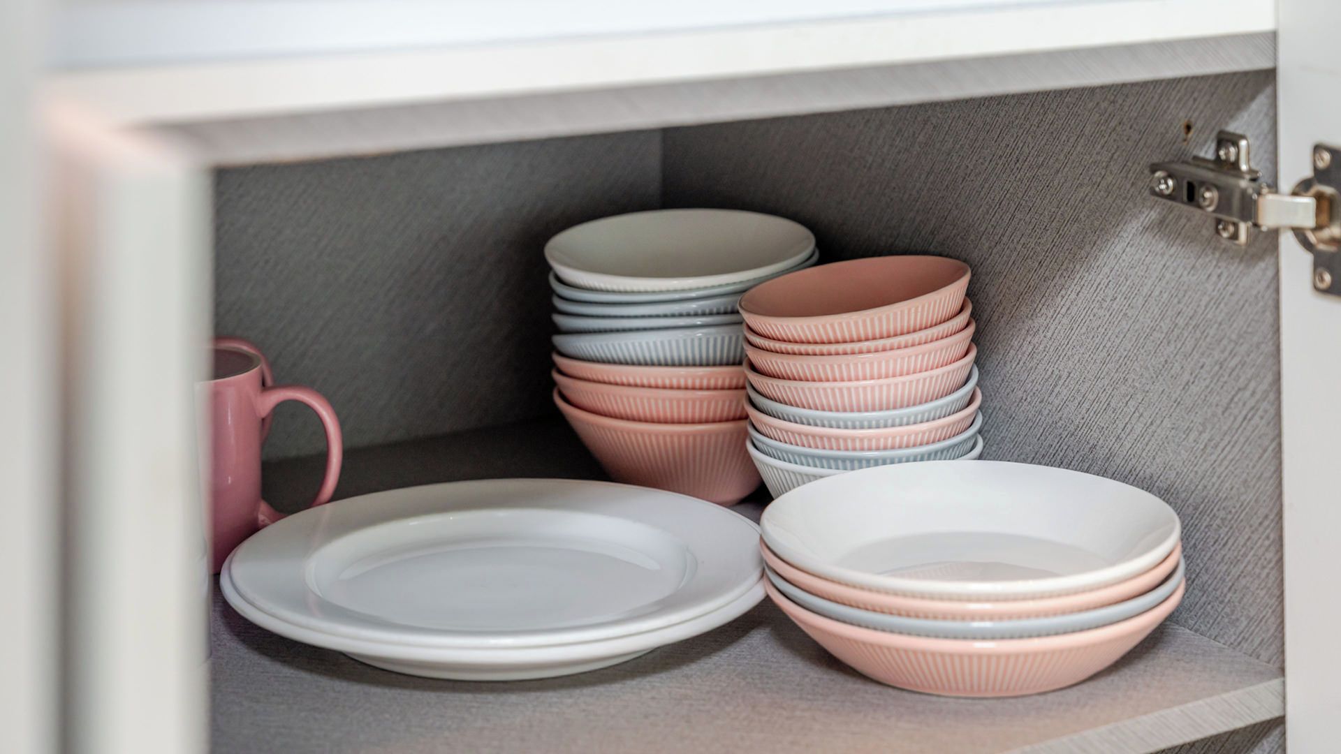 A cabinet with neatly stacked plates and bowls, a pink mug, and a metal latch.