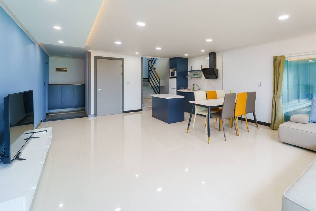 A modern living room with a white sofa, a dining table with chairs, a kitchen area, and a staircase leading to an upper floor.