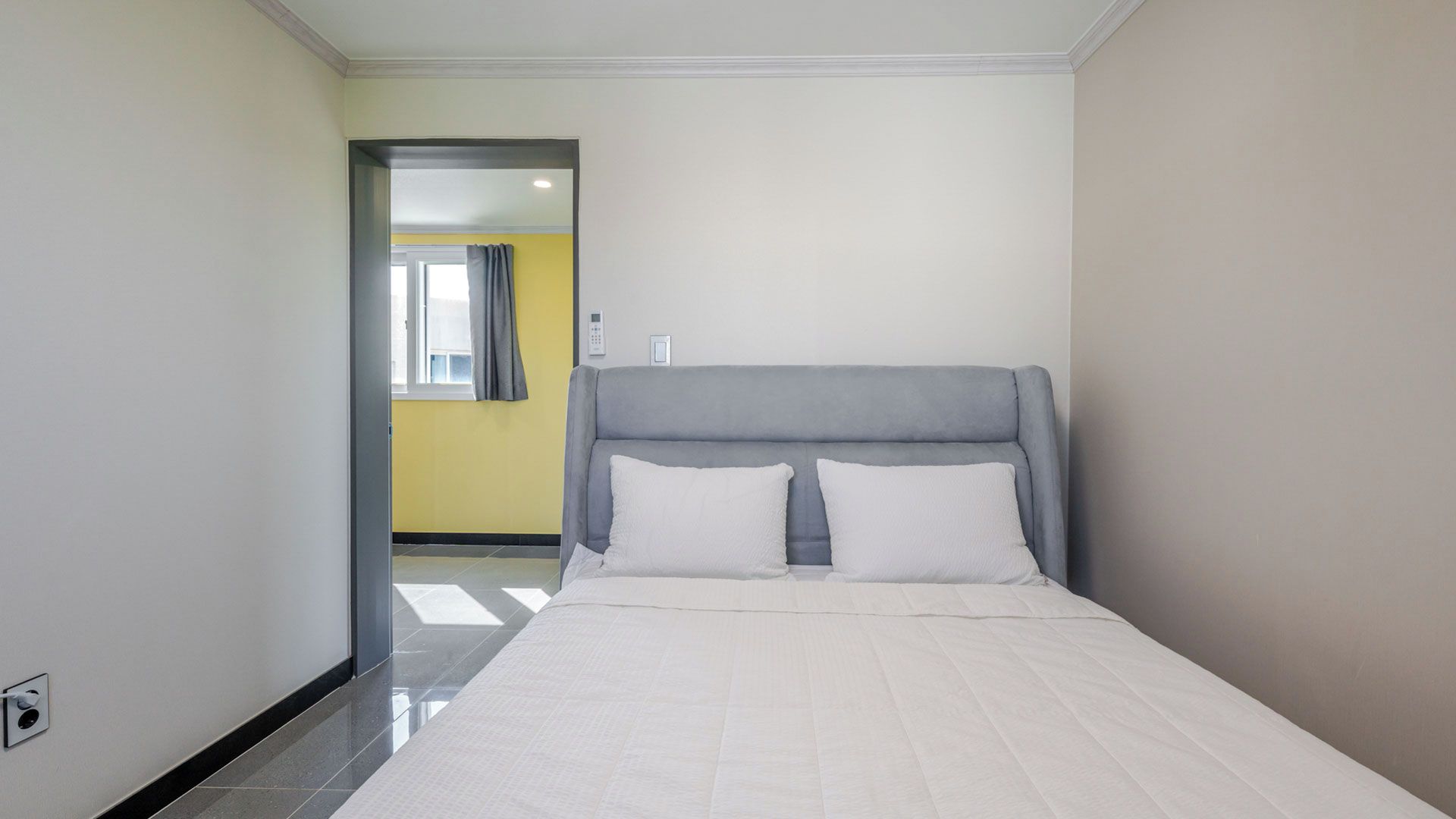 Ein Hotelzimmer mit einem Bett, einem Kopfteil, einem Fenster mit Vorhängen und einer Tür zu einem anderen Raum.