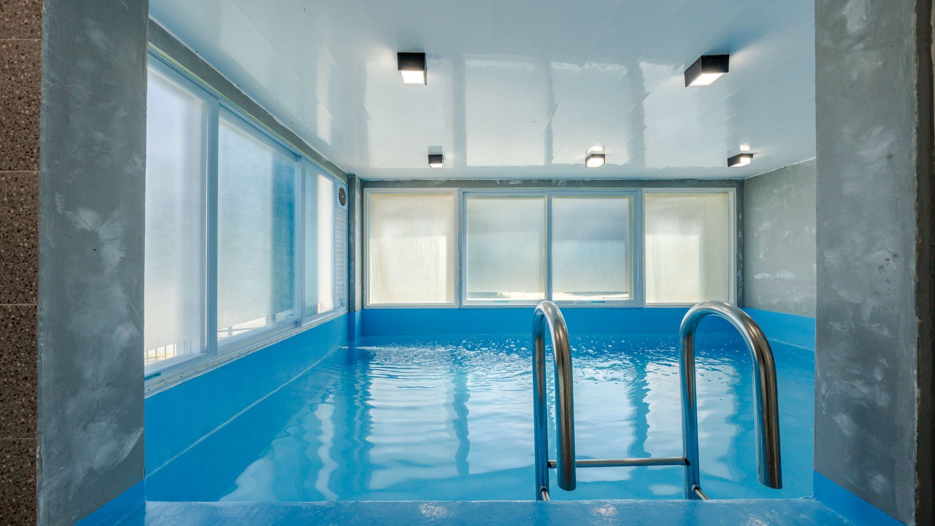 Une piscine intérieure avec des parois en verre transparent, un sol bleu et des échelles en métal.