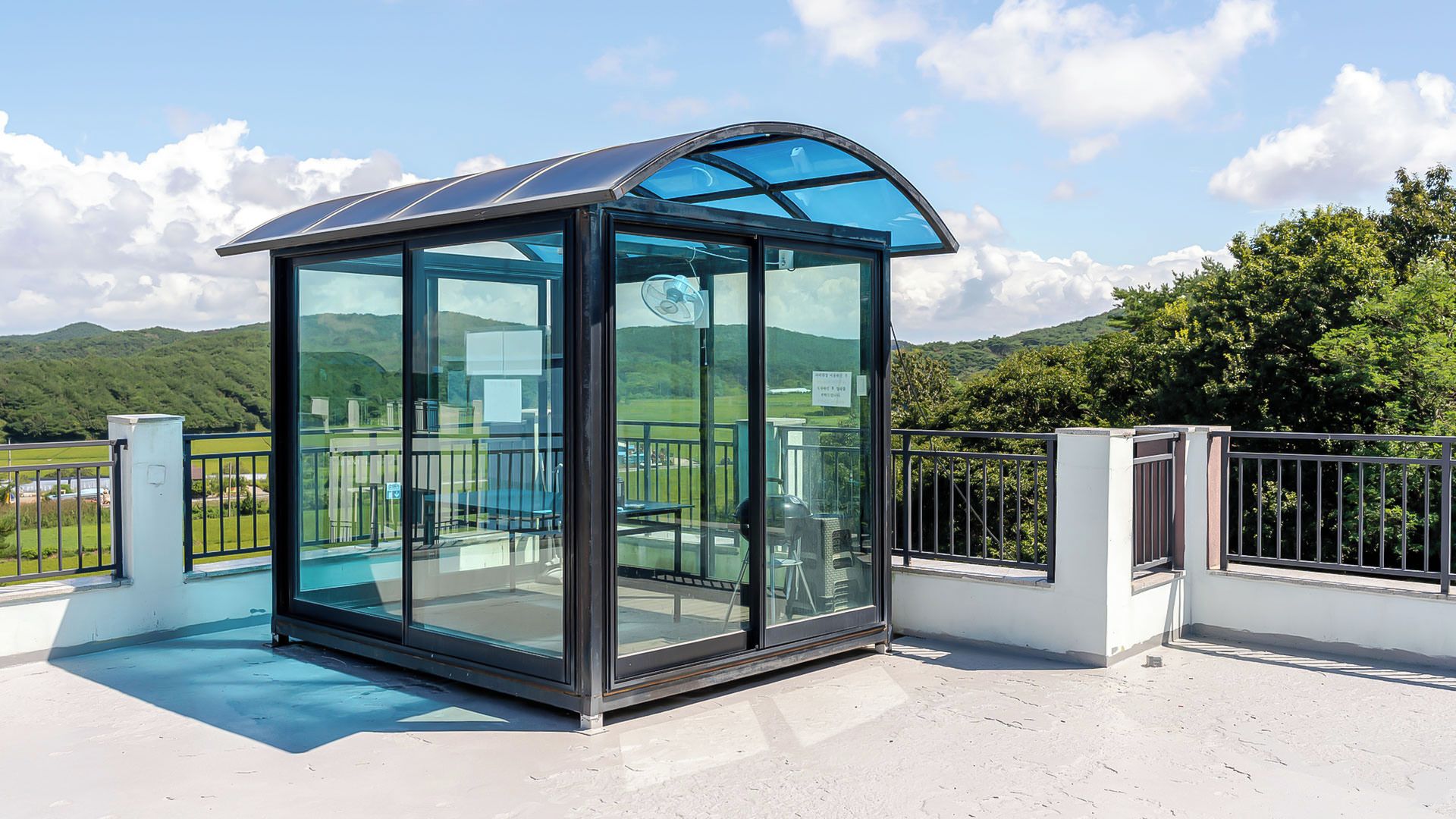 A glass-enclosed structure with a curved roof, a metal railing, and a view of a green landscape.