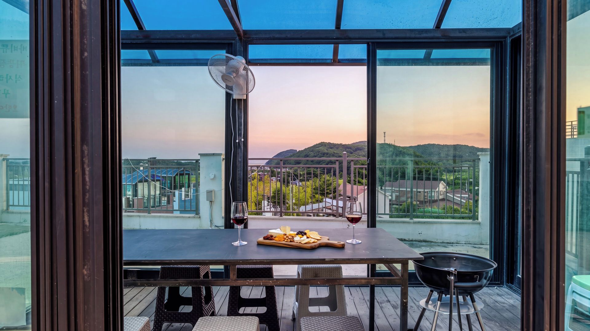 Un balcón con una mesa para dos, un ventilador y vistas a un paisaje urbano al atardecer.