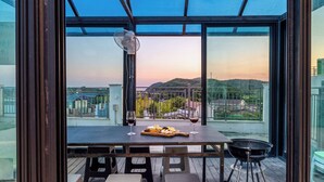 A balcony with a table set for two, a fan, and a view of a cityscape at sunset. - Taean Blueisland Pool Villa (Taean)