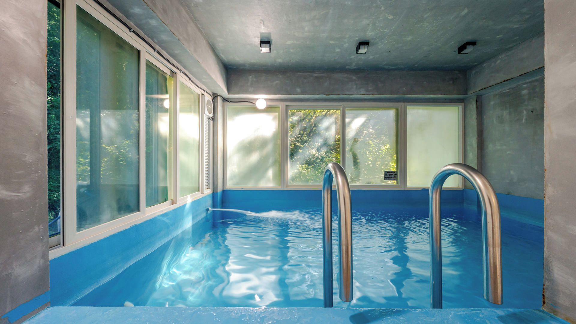 Una piscina interior con paredes de vidrio transparente, escaleras de metal y vistas a zonas verdes exteriores.