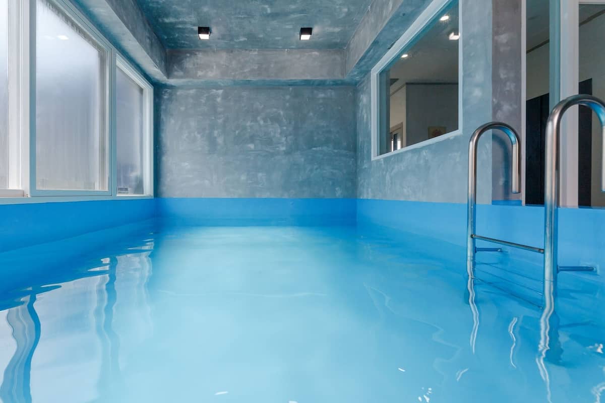 An indoor swimming pool with clear blue water, metal ladders, and a view of the surrounding area through large windows.