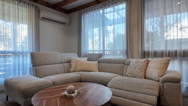 Um sofá modular com uma mesa de centro redonda, janelas amplas com cortinas leves e um aparelho de ar condicionado na parede.