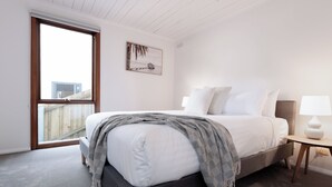 Um quarto com cama, mesa de cabeceira, abajur, janela com vista para edifícios e um quadro na parede.