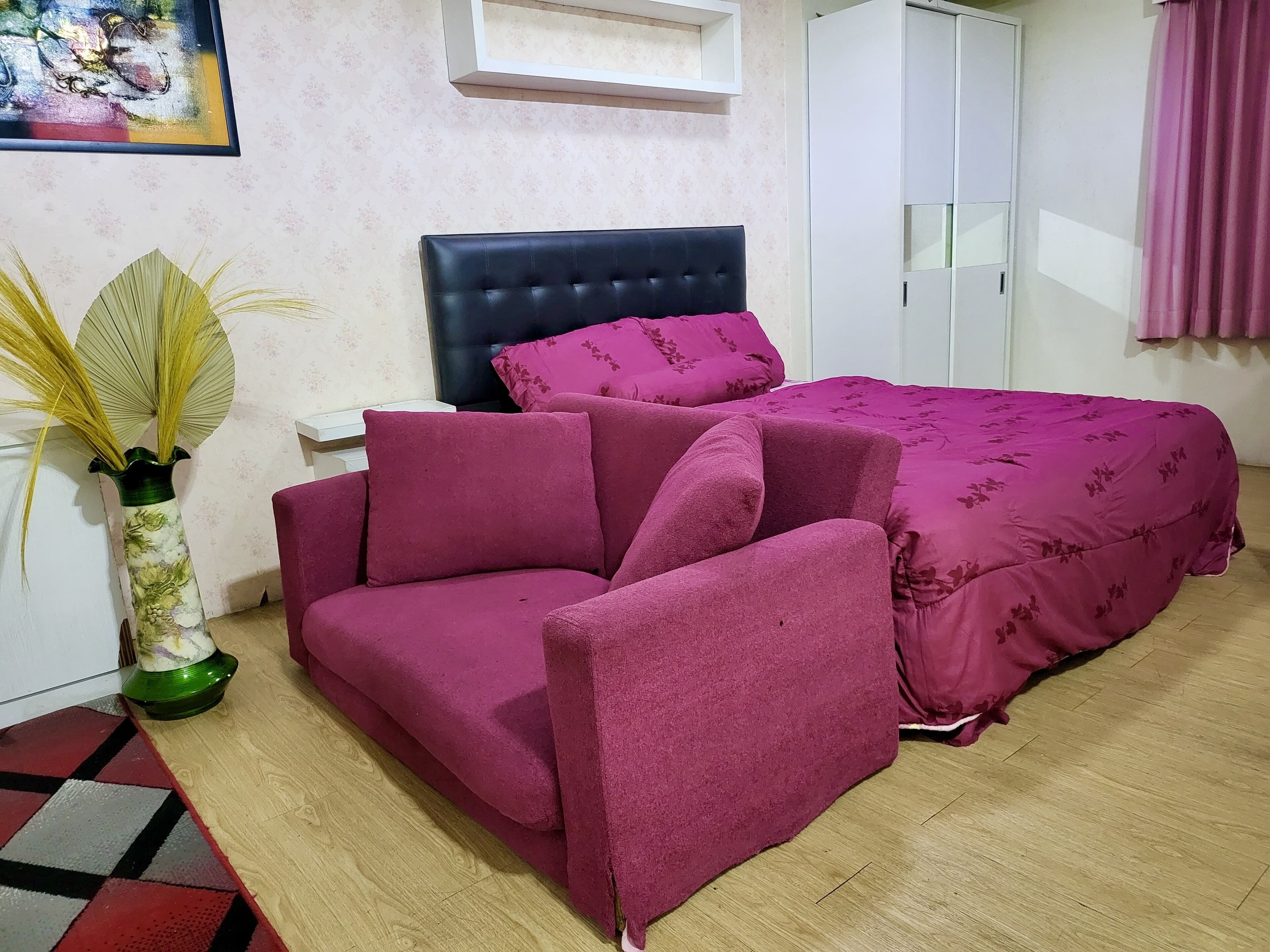A bedroom with a bed, a sofa, a nightstand, a vase with dried plants, and a painting on the wall.