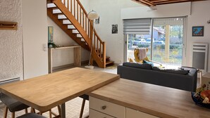 A modern interior with a wooden dining table, a sofa, a staircase, and a view through a sliding door.