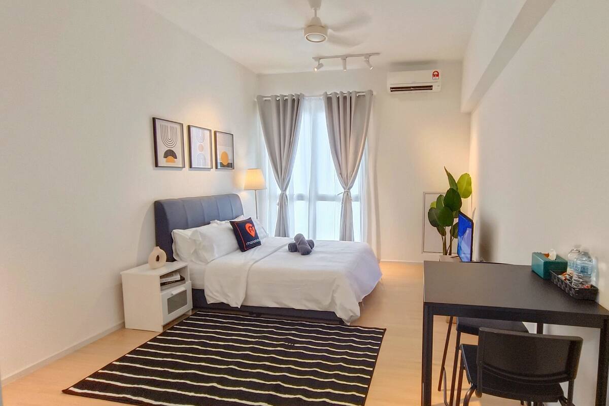 A hotel room with a bed, bedside table, desk, chair, and a large window with curtains.