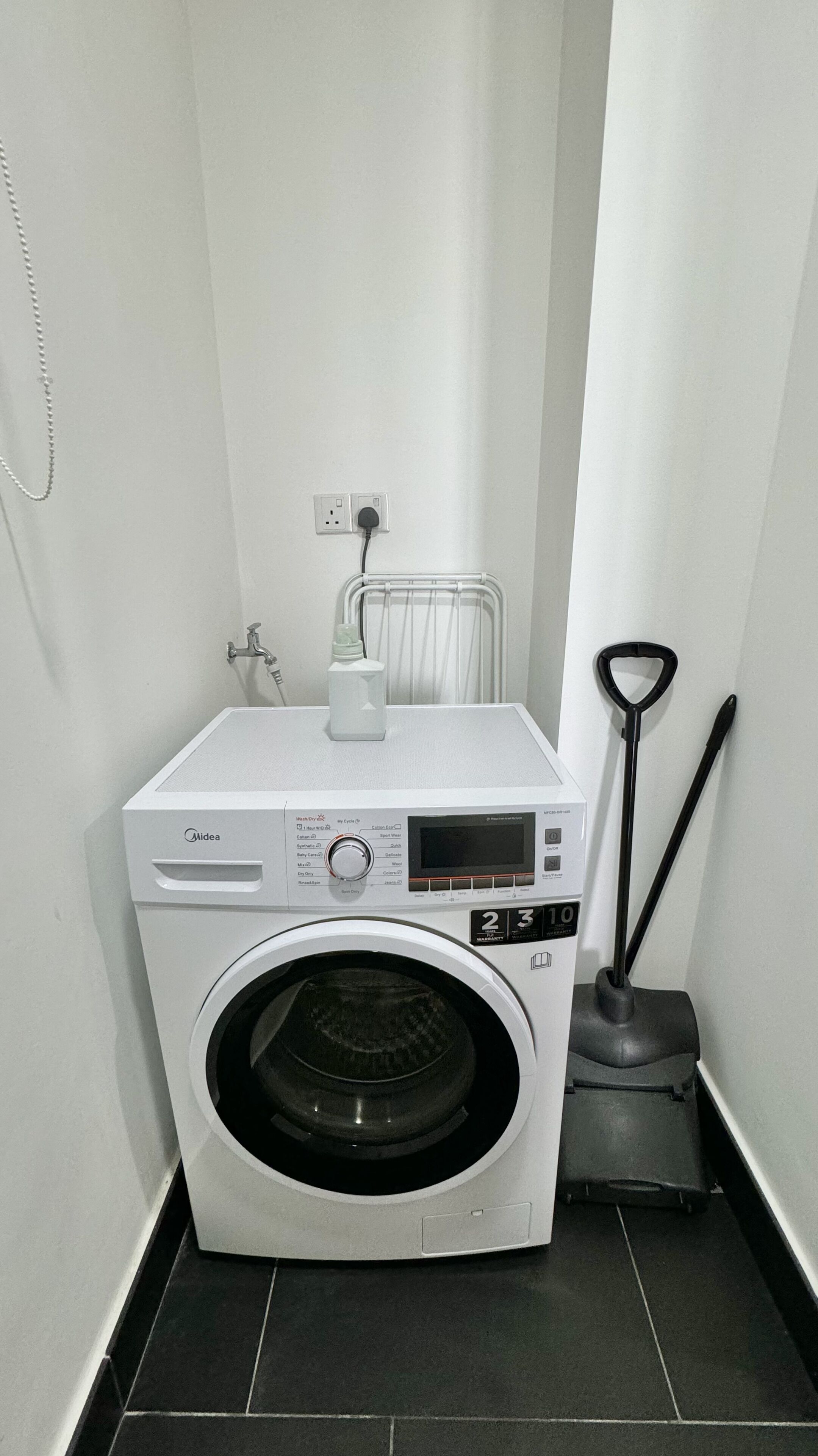 A compact, white Midea washing machine in a small laundry room with a tiled floor.