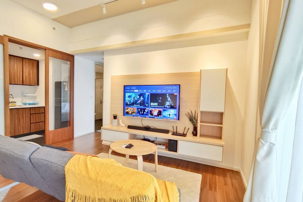 A modern living room with a grey sofa, a yellow throw blanket, a flat-screen TV mounted on the wall, a small wooden coffee table, and a built-in kitchen area with wooden cabinets.