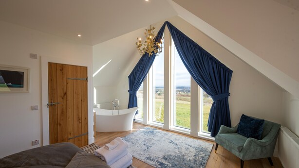 A bedroom with a large window, a bathtub, a green armchair, and a wooden door. - Robin Cottage, Logiealmond Estate (Perth)