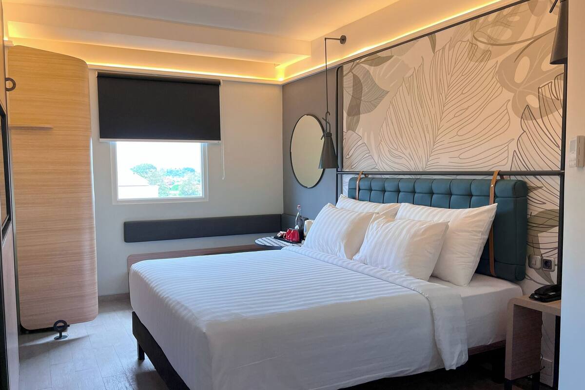 A modern bedroom with a large bed, a bedside table, a mirror, and a window with a view of greenery.