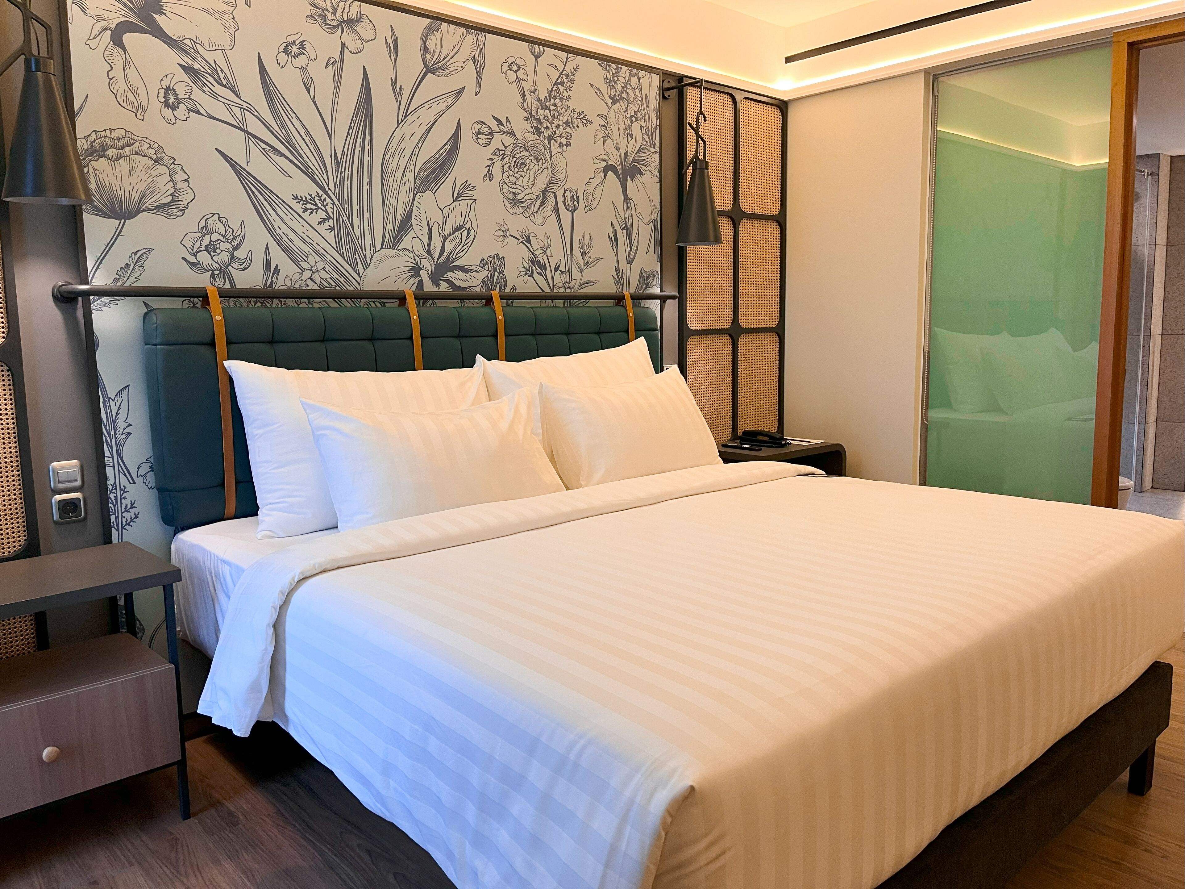 A neatly made bed with white linens, a green upholstered headboard, and a wall with a floral pattern.