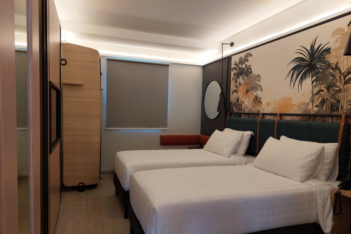 A hotel room with two beds, a large window, a wall-mounted TV, and a tropical-themed mural.