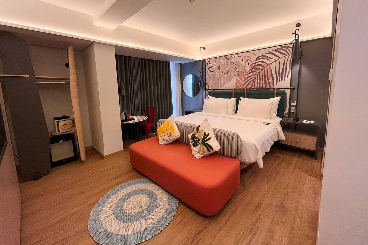 A modern hotel room with a large bed, a red ottoman, a round rug, a desk with a chair, and a view of the city through the window.