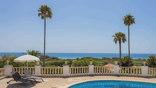 A pool area with two lounge chairs and an umbrella, surrounded by a white fence and palm trees, overlooking a beach. - Villa Bautista - Beautiful Sea View (San Jaime Mediterráneo)