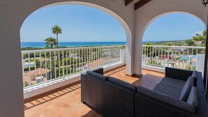 A balcony with a wicker sofa and a white railing, overlooking a coastal landscape with palm trees and a clear sky. - Villa Bautista - Beautiful Sea View (San Jaime Mediterráneo)