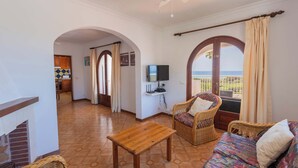 A room with a wooden table, wicker furniture, a sofa, a television, and a view of the sea through an arched window. - Villa Bautista - Beautiful Sea View (San Jaime Mediterráneo)