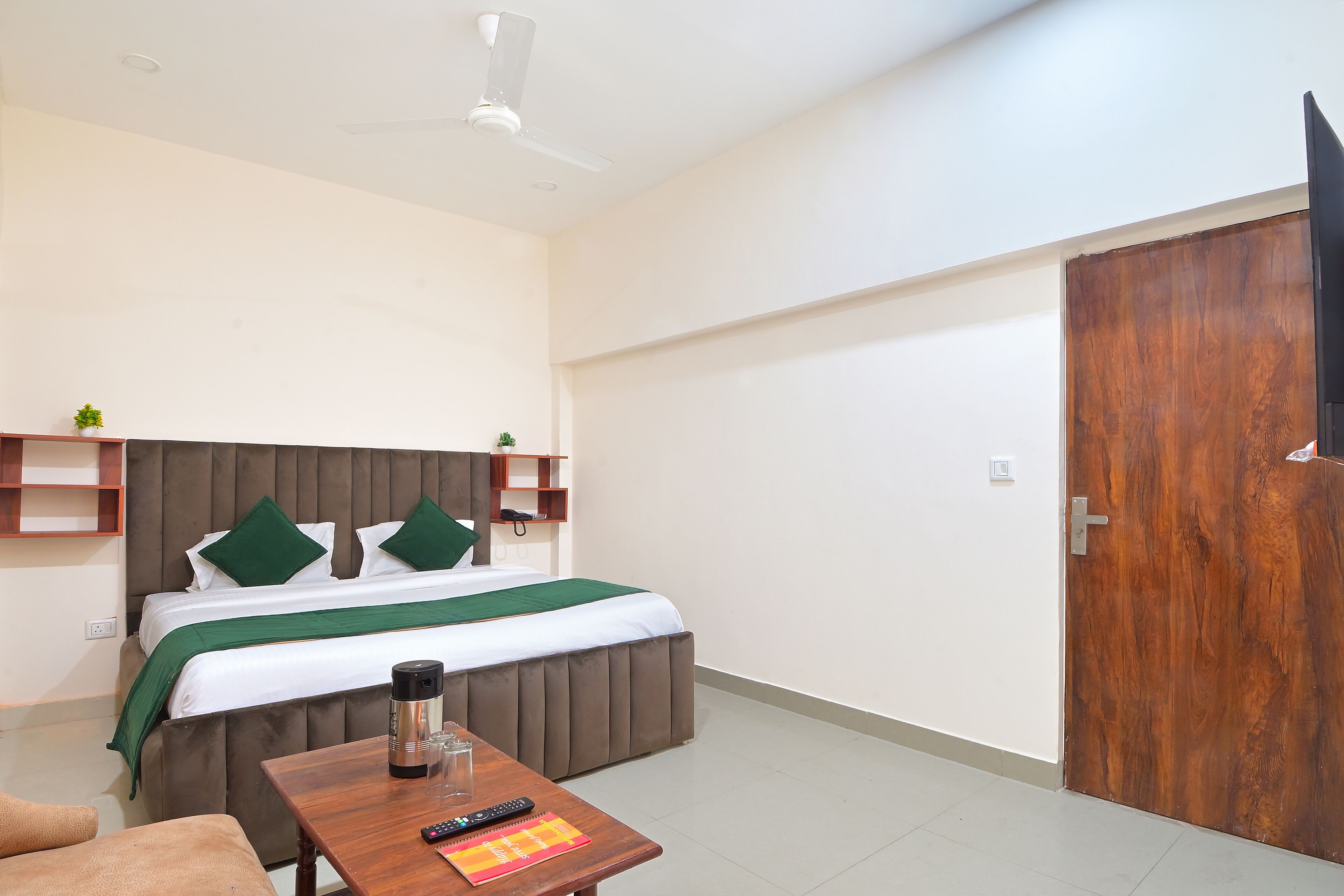 Habitación de hotel con cama, una mesa de madera con un control remoto, cafetera y televisor en la pared.