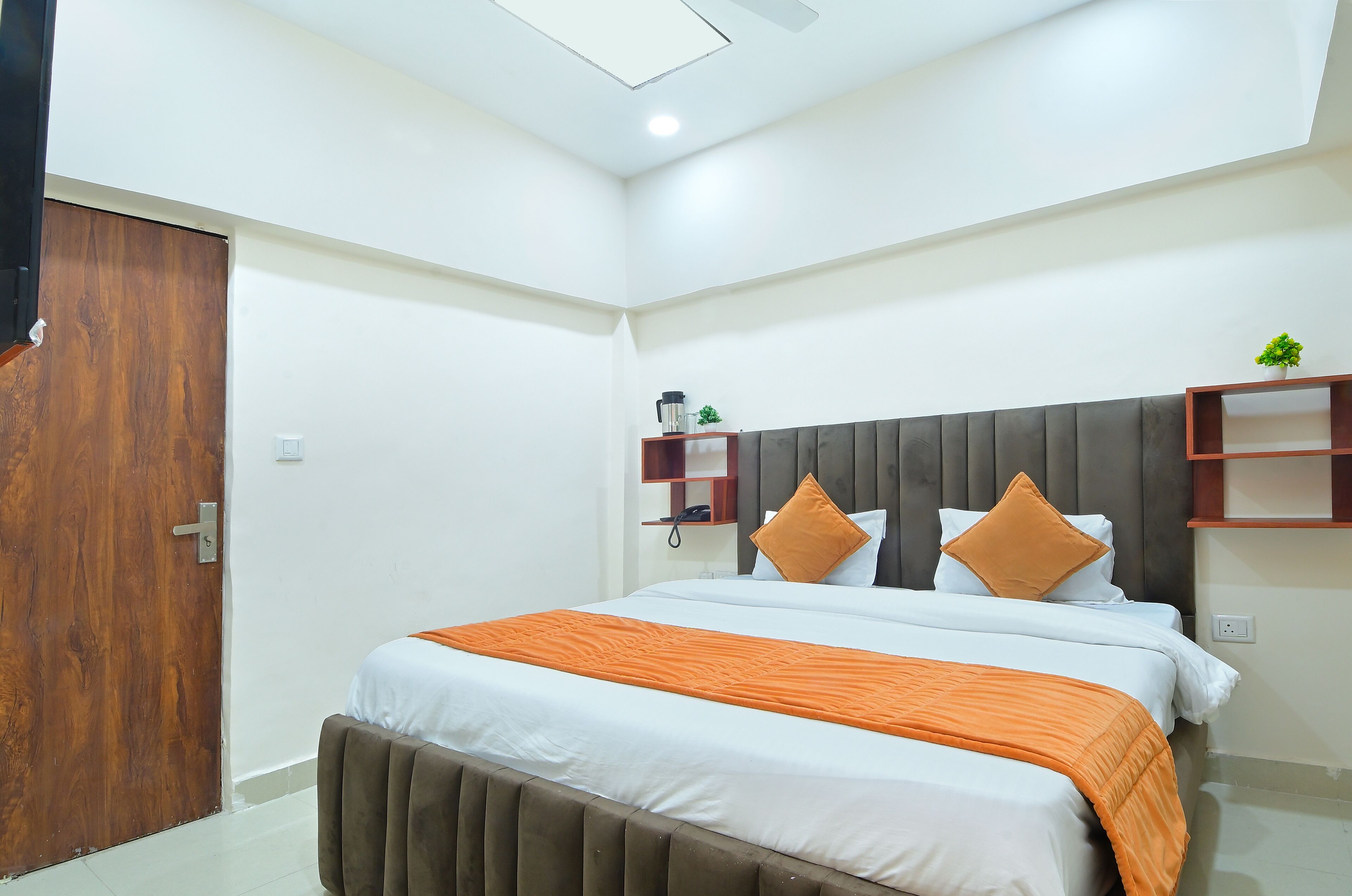 A modern hotel room with a large bed, a wooden door, a small plant, and a shelf with a phone and a coffee maker.