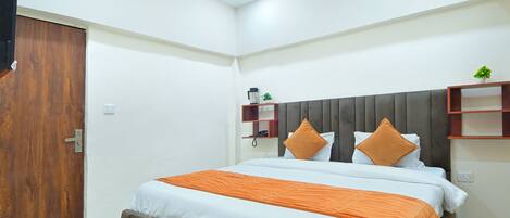 A modern hotel room with a large bed, a wooden door, a small plant, and a shelf with a phone and a coffee maker.