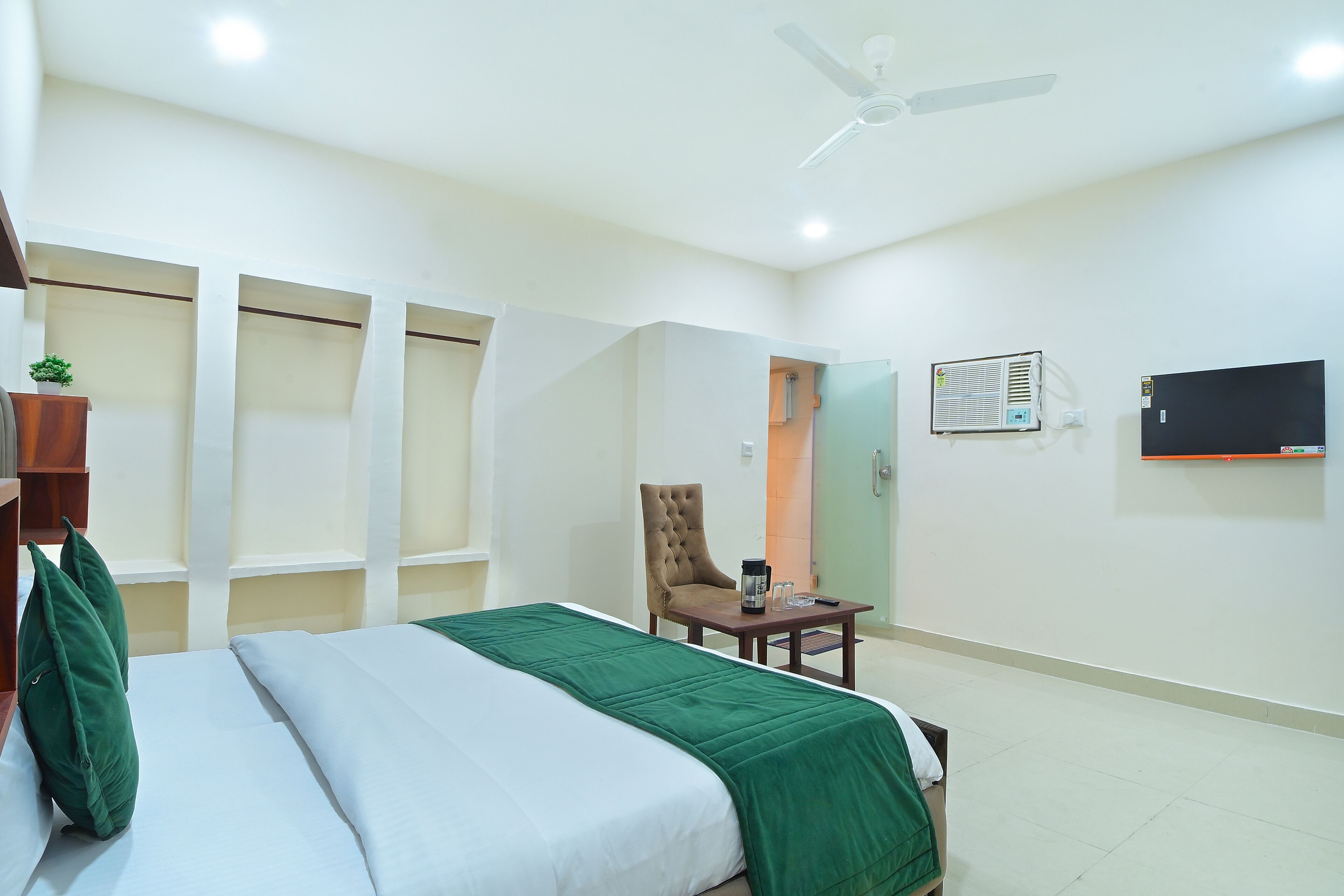 Una habitación de hotel con cama, una silla, una mesa con cafetera, un equipo de aire acondicionado y un televisor en la pared.