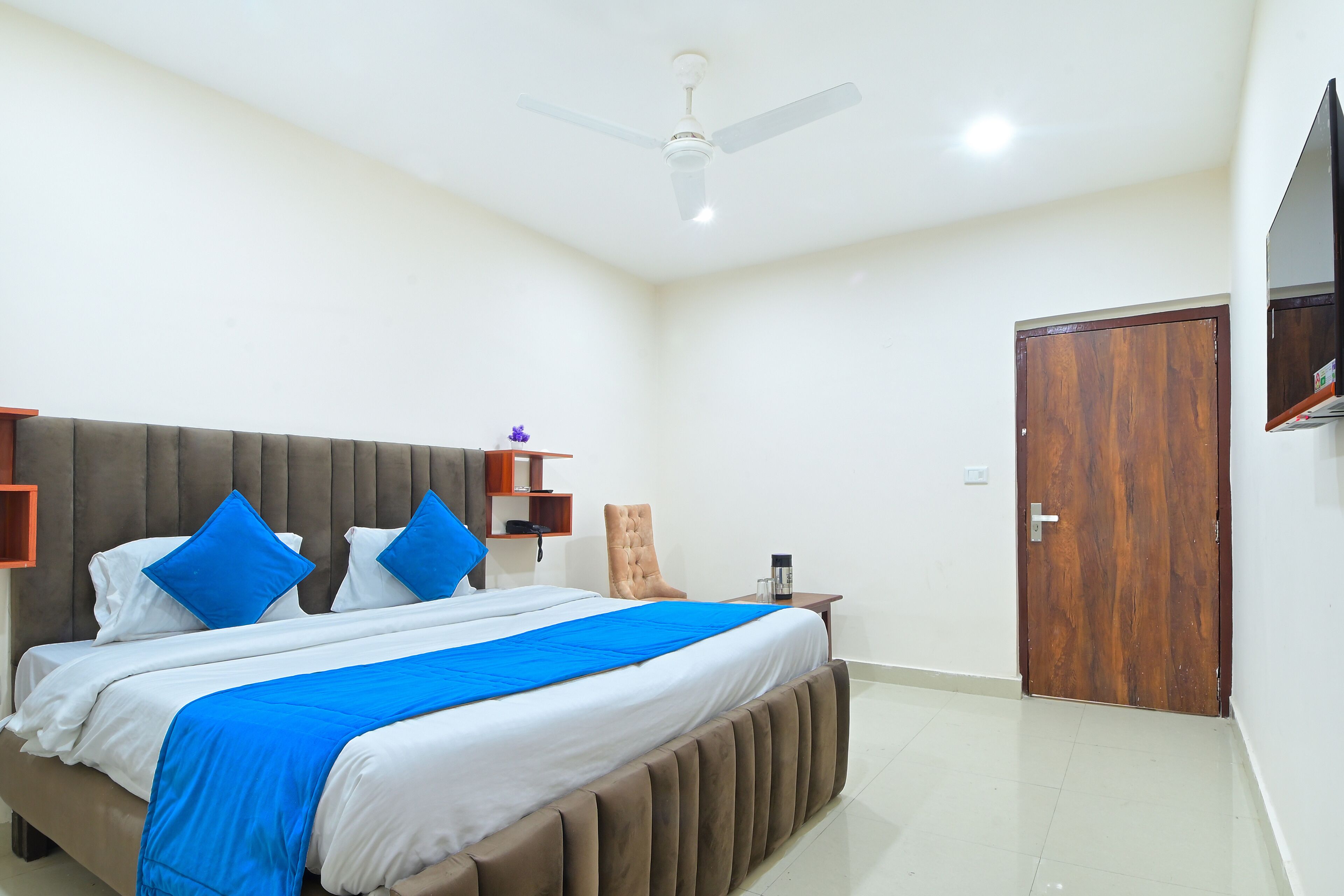 Habitación de hotel con una cama grande, un ventilador de techo, una puerta de madera y un televisor montado en la pared.
