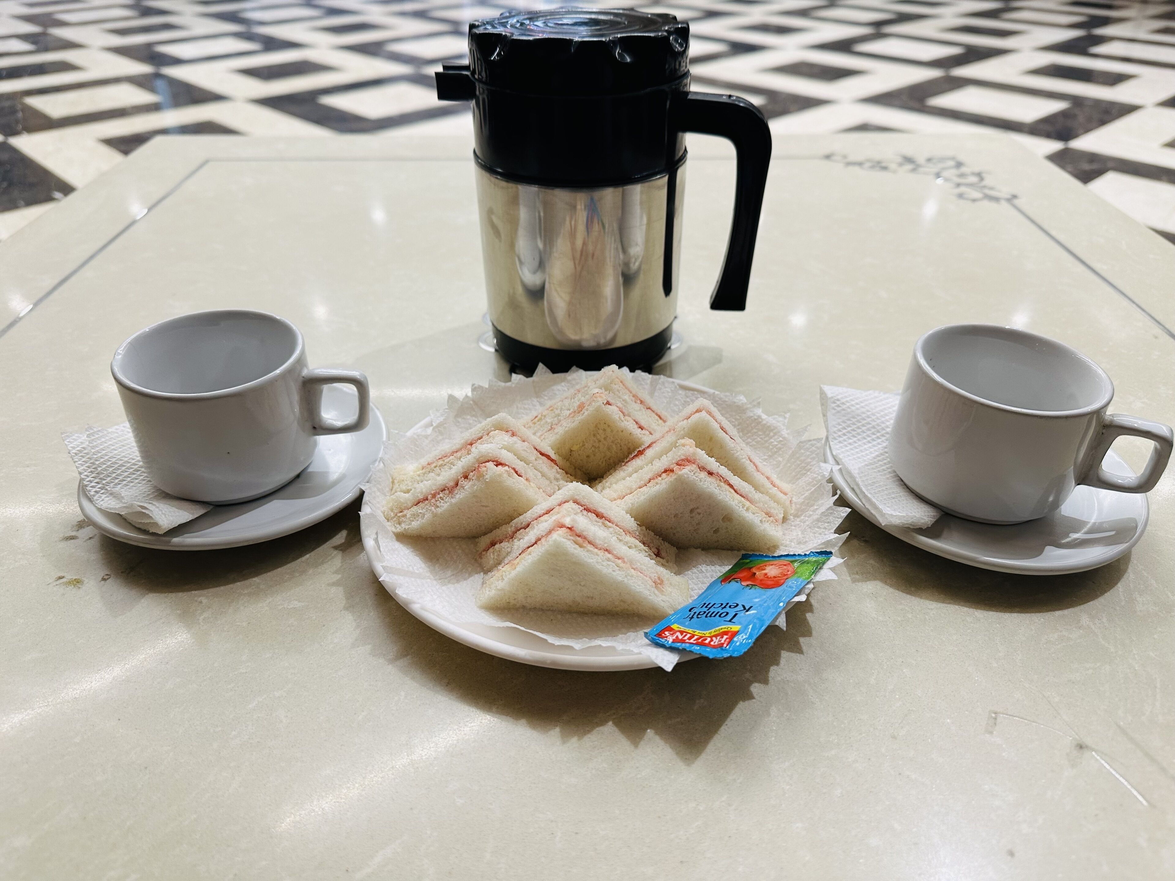 A table with a plate of sandwiches, two cups, and a coffee maker.