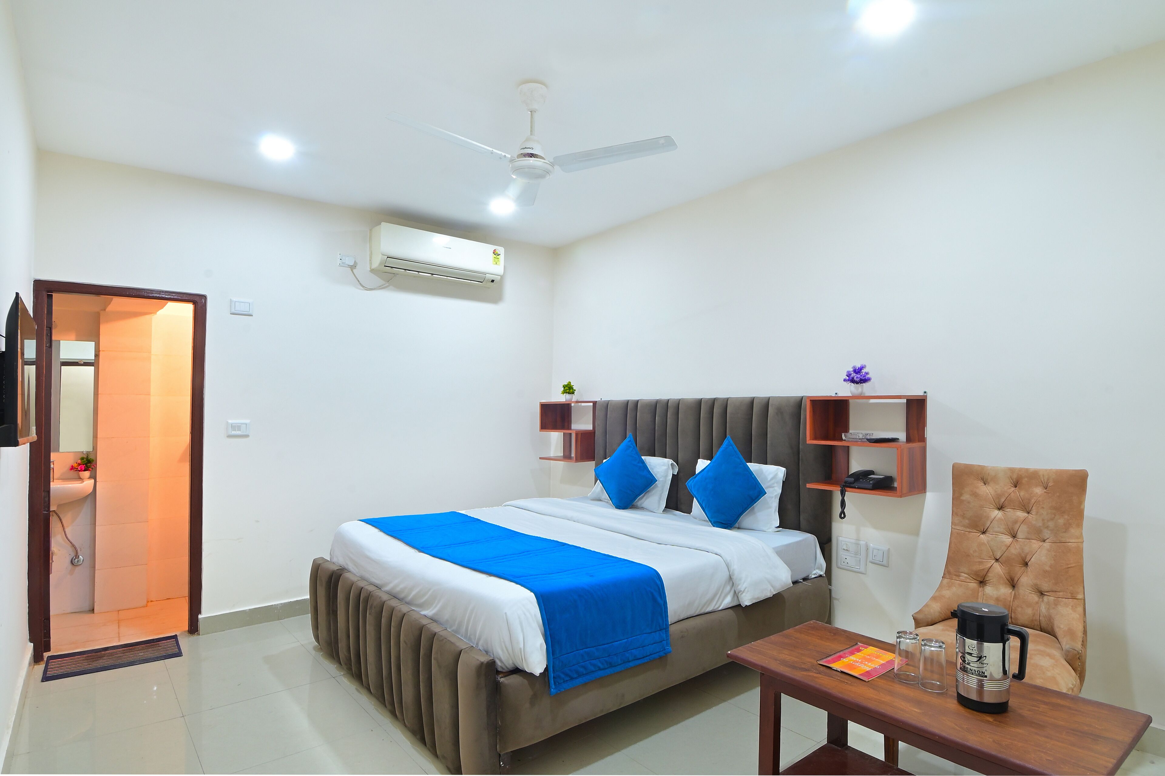 A hotel room with a bed, a chair, a table, a ceiling fan, an air conditioning unit, and a small shelf with a plant.