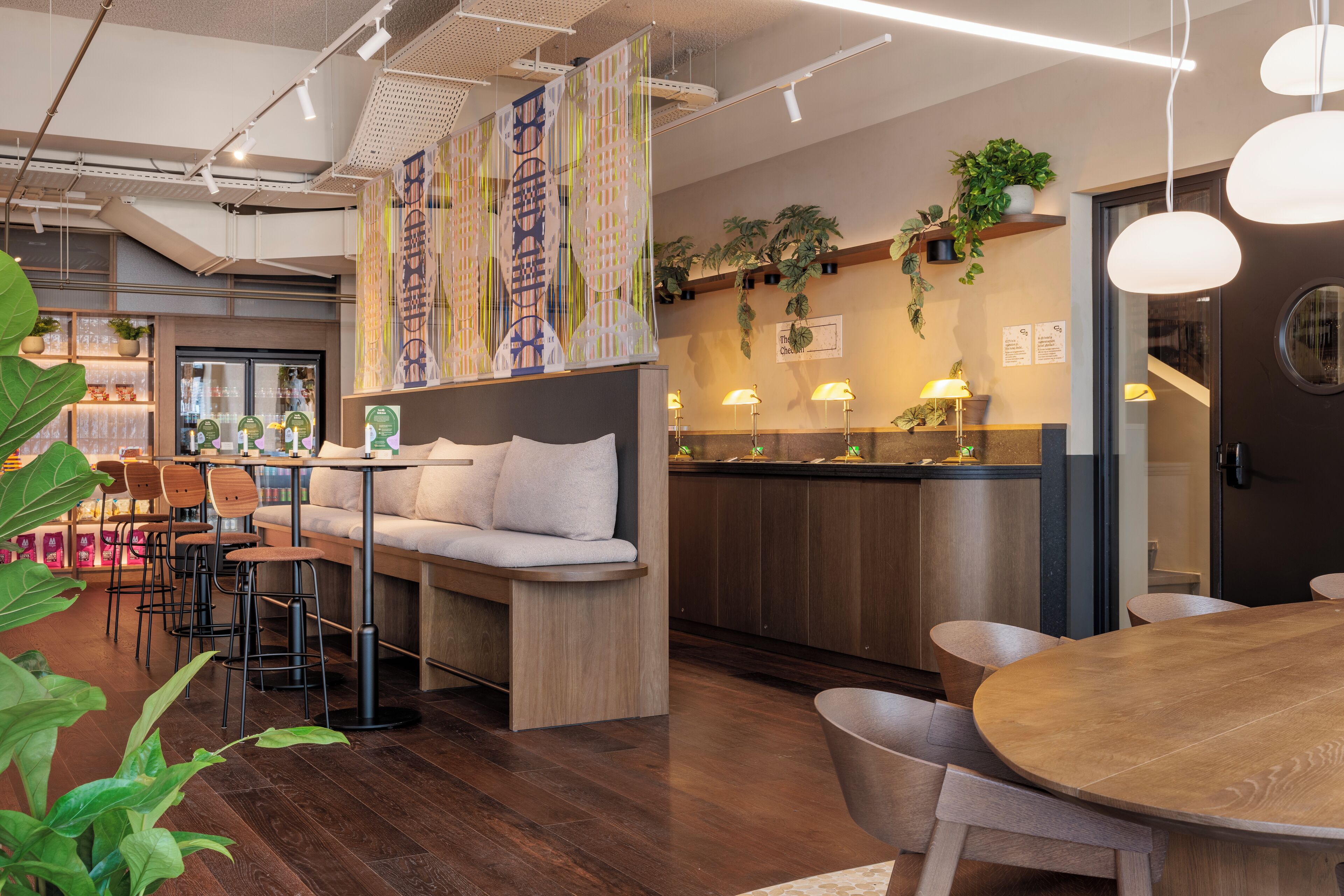 A modern interior with a wooden dining table, cushioned seating, and a kitchen area with plants and lighting.