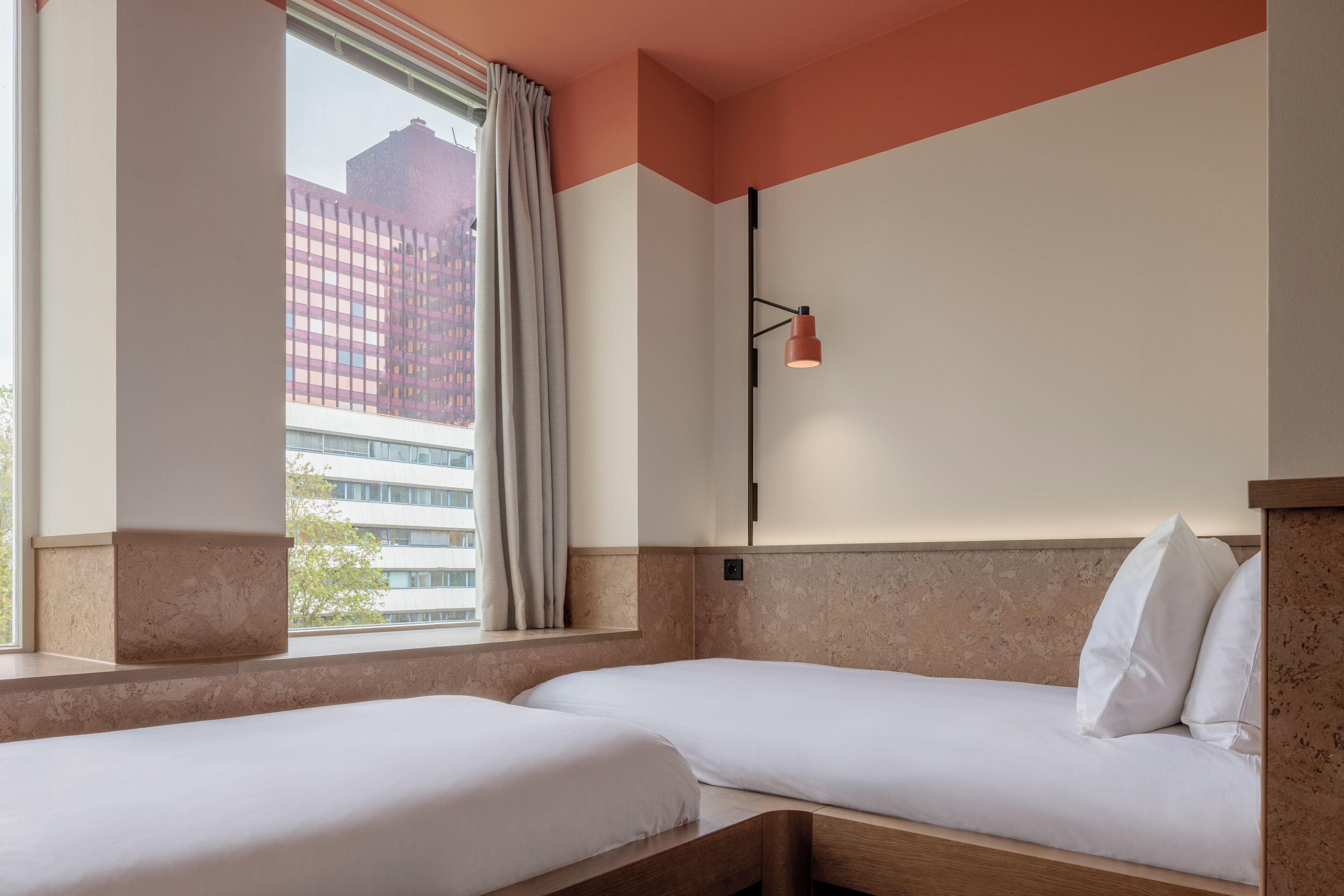 Habitación de hotel con dos camas, ventana con cortinas, lámpara de pared y vista a un edificio.