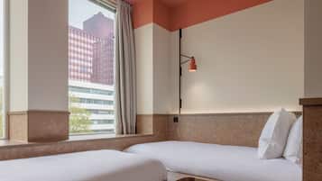 A hotel room with two beds, a window with curtains, a wall-mounted lamp, and a view of a building outside.