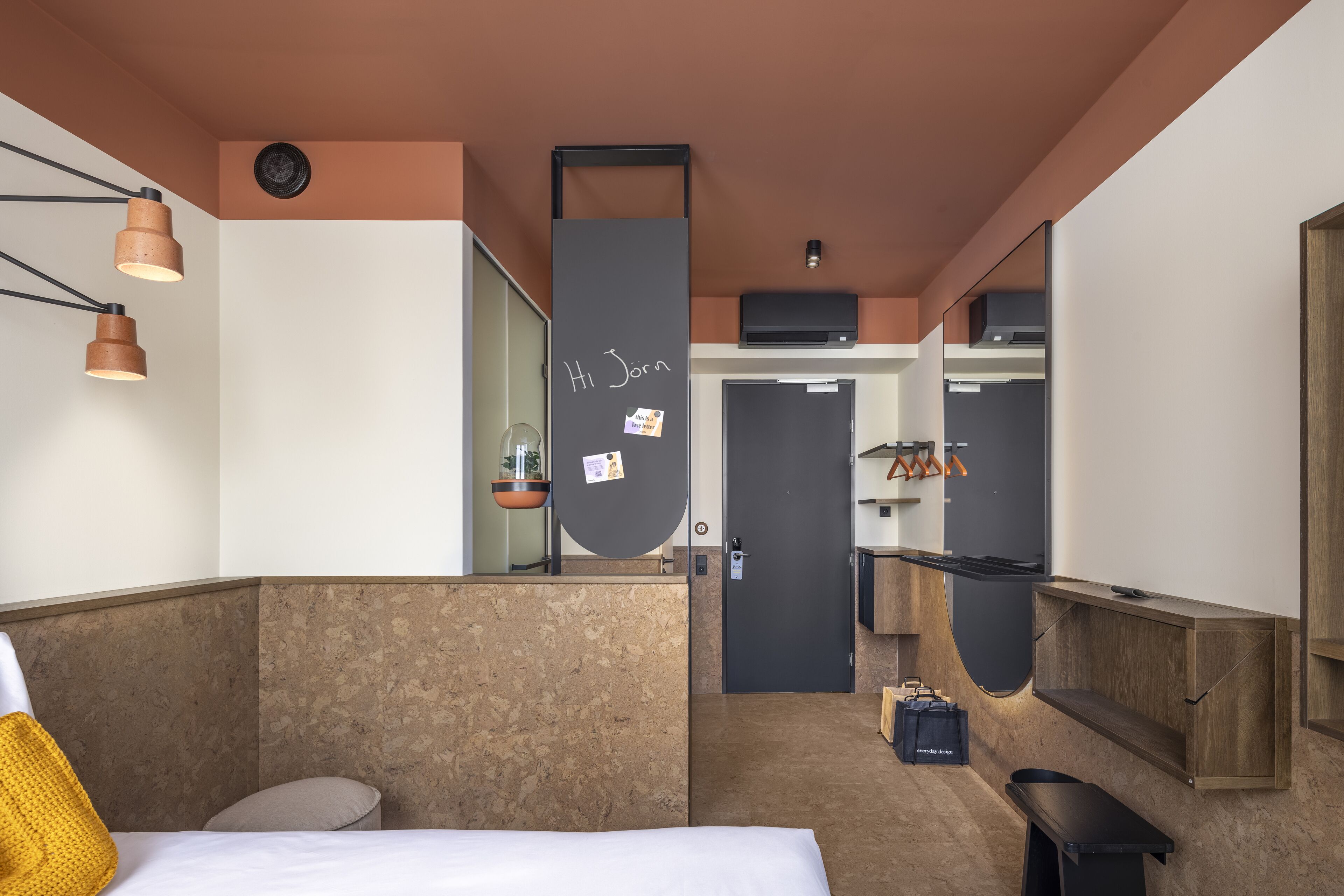 A modern hotel room with a bed, a desk, a mirror, and a small bathroom.