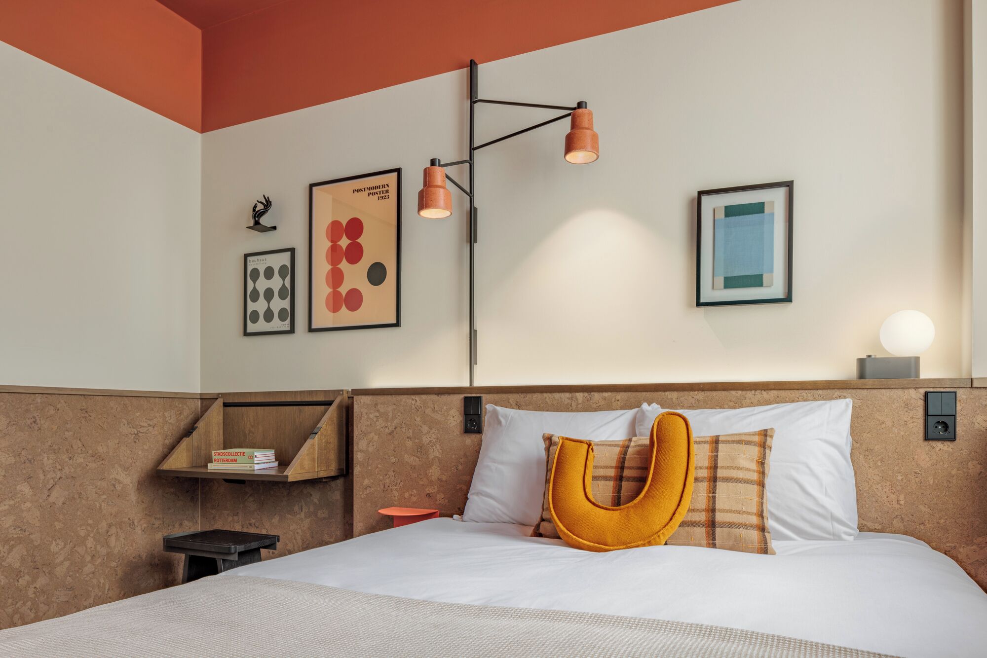 Una habitación de hotel con cama, mesita de noche, estante con libros y obras de arte en la pared.