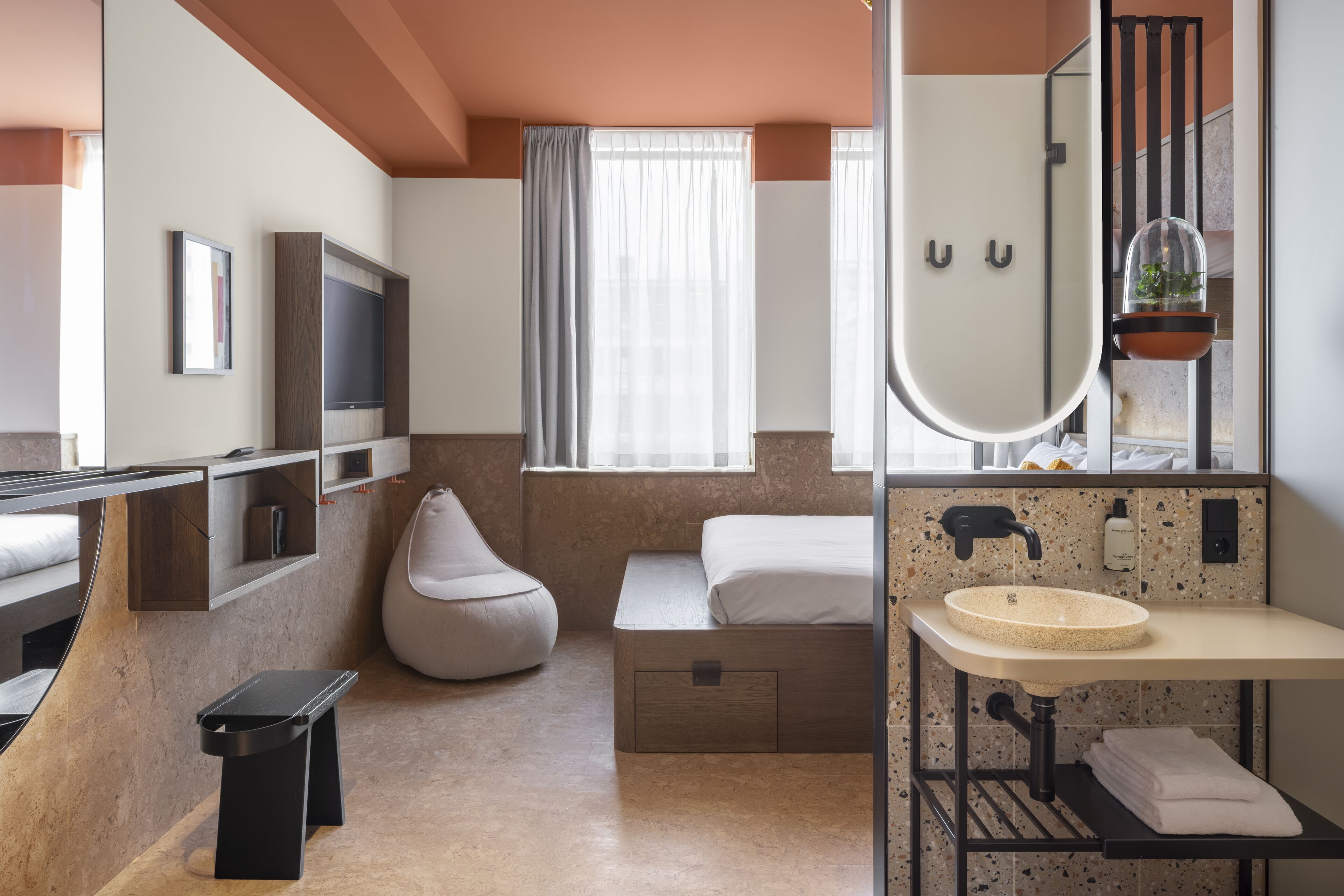 A modern hotel room with a curved bed, a built-in TV, a large mirror, a sink, and a potted plant.