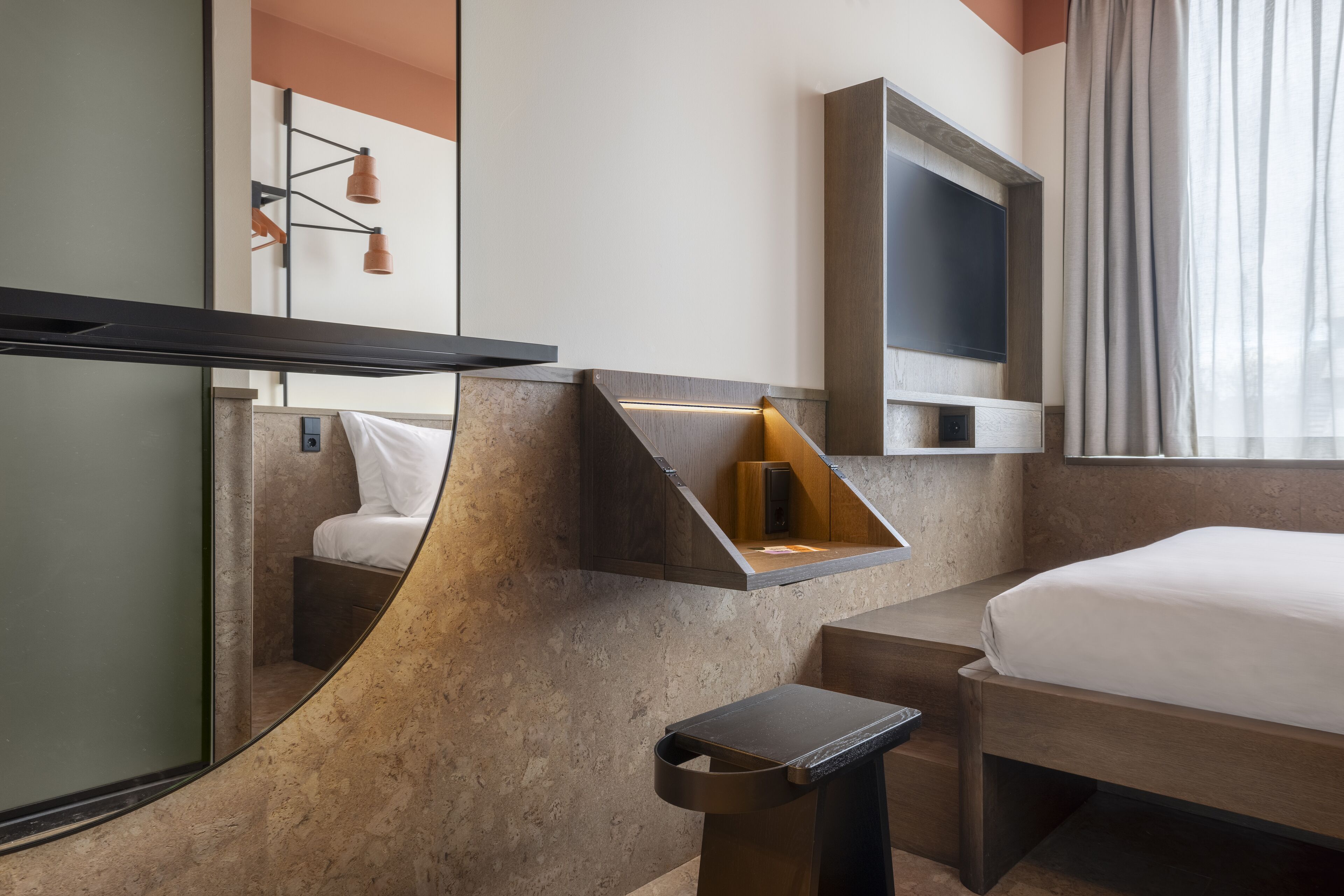 A modern hotel room with a bed, a small desk, a mirror, and a wall-mounted TV.