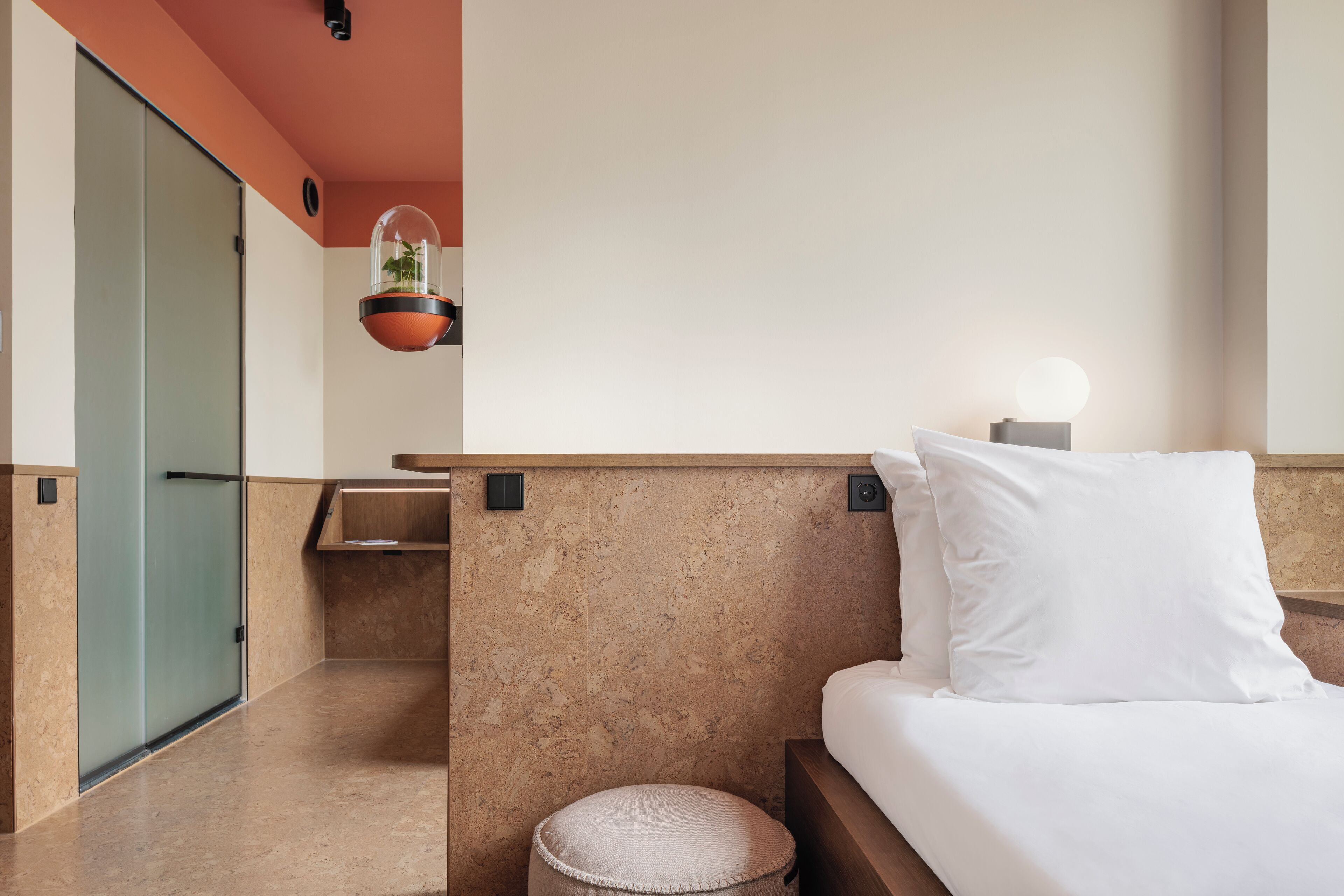 Ein Hotelzimmer mit Bett, Nachttisch, hängender Pflanze und kleinem Hocker.