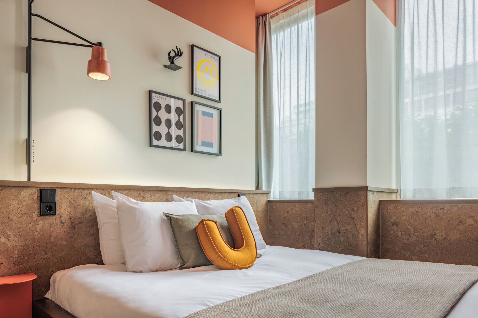 A modern hotel room with a bed, a nightstand, a wall-mounted lamp, and framed artwork.
