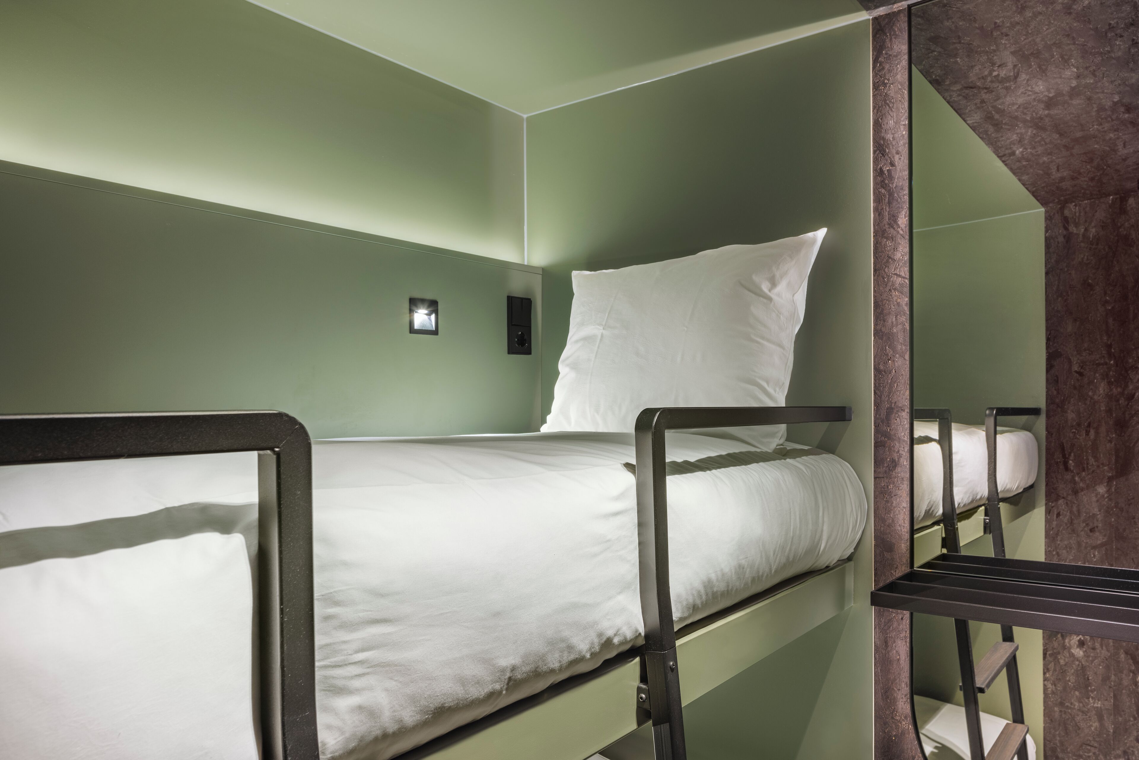 Ein Stockbett mit weißer Matratze und Kissen, einer kleinen Wandleuchte und einem dunkel gefärbten Metallrahmen.