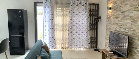 A modern living room with a sofa, a TV on a wooden stand, a black refrigerator, and a patterned curtain.
