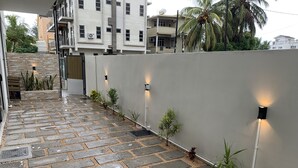 Un camino pavimentado con plantas pequeñas y luces fijadas en la pared, que conduce a un edificio con varios balcones.