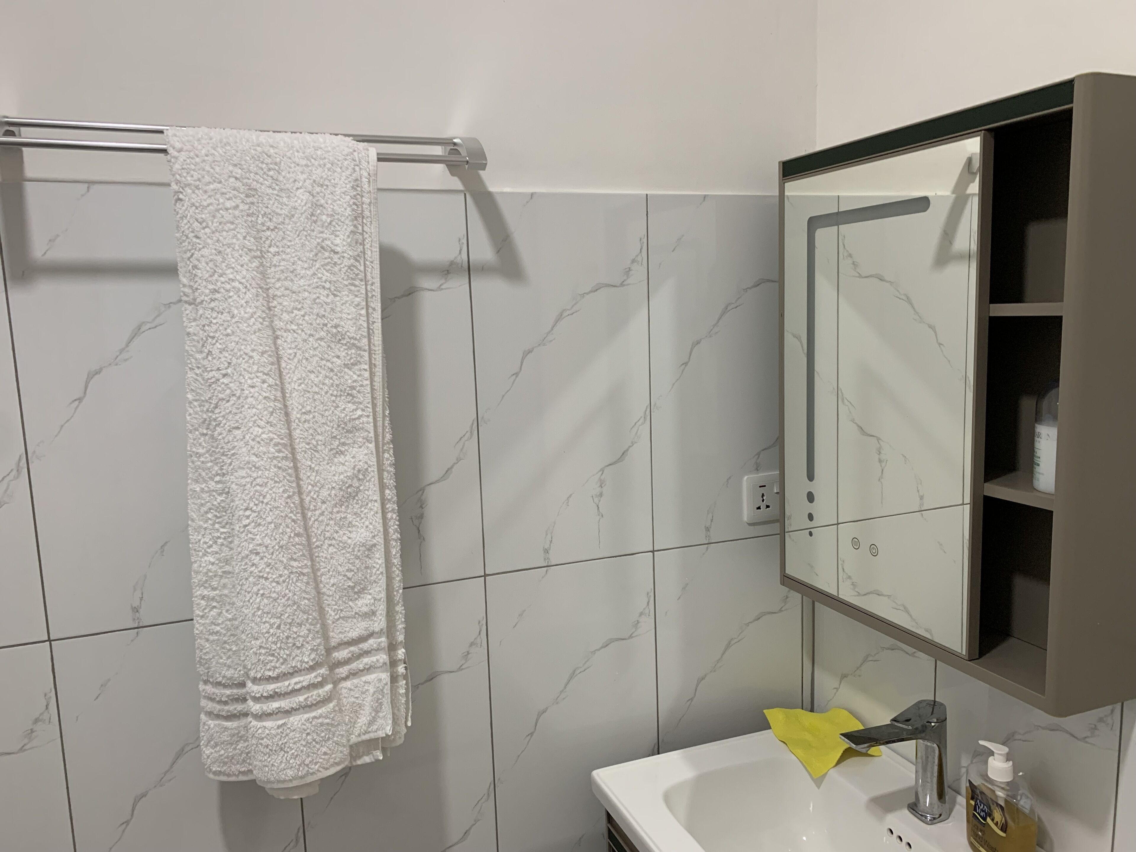 A bathroom with a white marble tiled wall, a white towel hanging on a metal towel rack, a white sink with a yellow cloth, and a mirror with a shelf above it.
