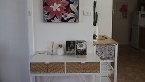 Et hvidt konsolbord med et blomstermotiv over sig, en kaktus og en lille plantekasse. Der er to dekorative æsker og et skilt med teksten "Laugh" på bordet.