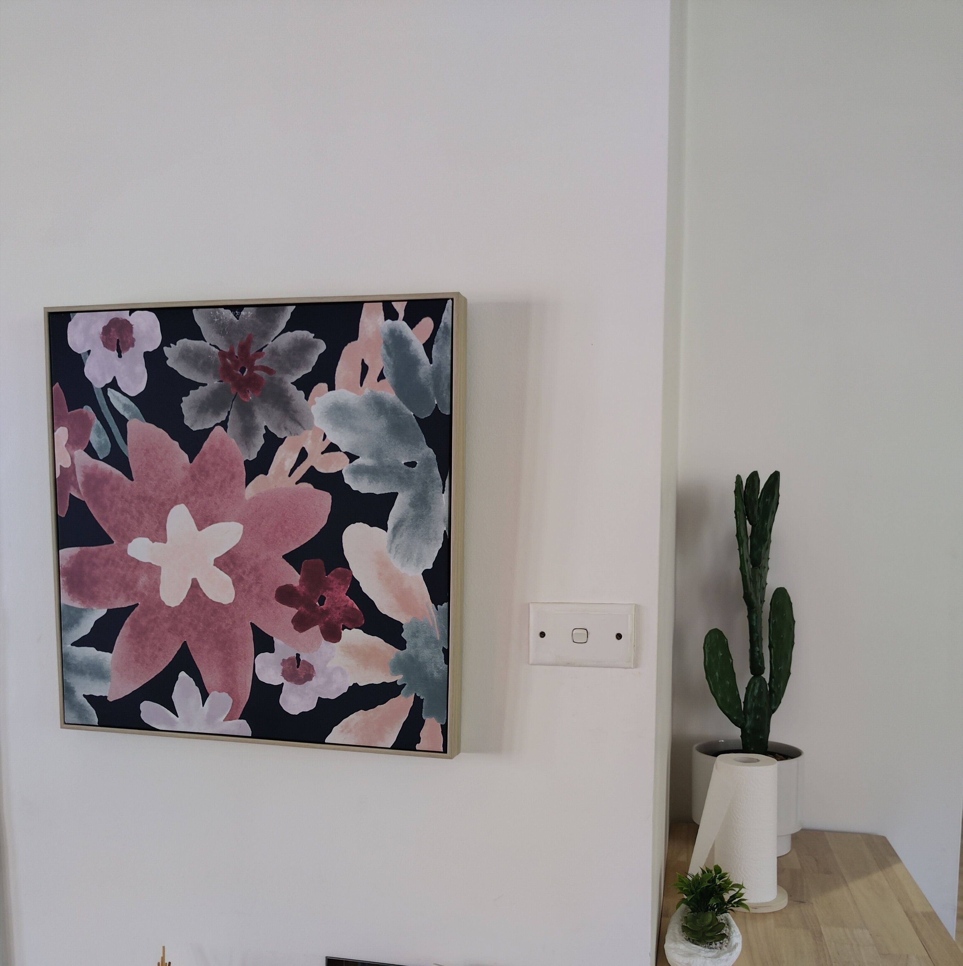 Una obra d'art floral emmarcada a una paret blanca, una presa elèctrica blanca i dues plantes en testos sobre una superfície de fusta.