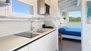 Una cocina moderna con ventana, un lavamanos y una encimera. En la habitación contigua, hay una cama con colcha azul.