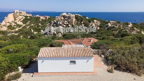 Un edificio blanco con tejado de baldosas, rodeado de terreno rocoso y vegetación, con vistas a la costa.